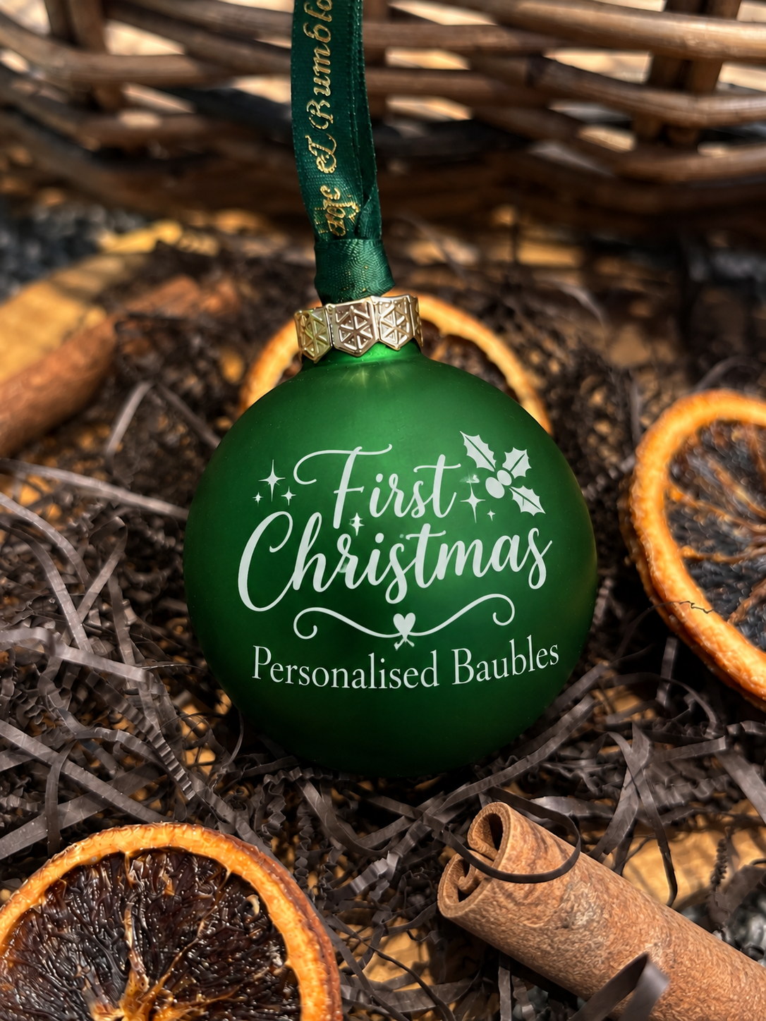 A green Christmas ornament with 'First Christmas Personalised Baubles' printed in white, hanging from a dark green ribbon with gold lettering. The ornament is styled in a rustic setting with dried orange slices, a cinnamon stick, and shredded paper.