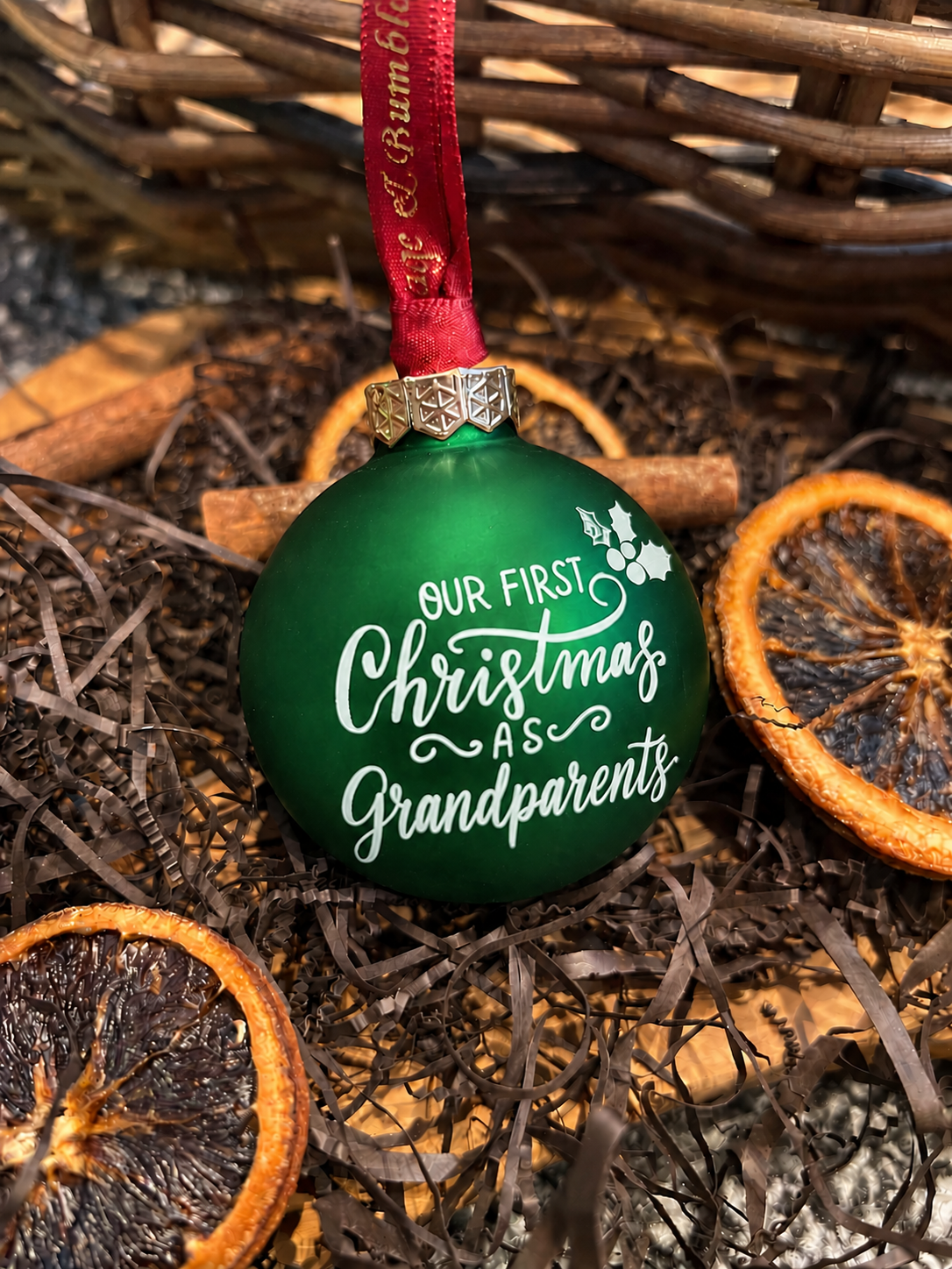 A green Christmas ornament with white script text reading 'OUR FIRST Christmas AS Grandparents' hanging from a red ribbon, styled in a basket with dried orange slices and cinnamon sticks.