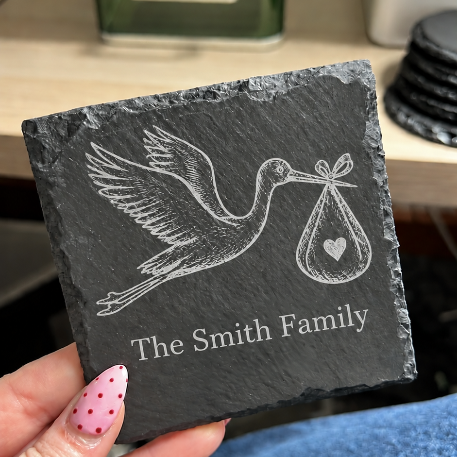 A hand holds a square dark slate coaster engraved with a stork carrying a baby bag and the text 'The Smith Family'. The background shows a desk with a green bottle and other office items, along with blue jeans.